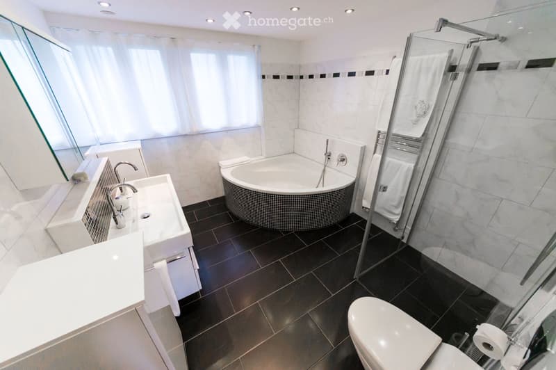 Modern bathroom with a white sink, bathtub, toilet, and glass-enclosed shower. Features include a large window with blinds, white tiles, and a dark tiled floor.