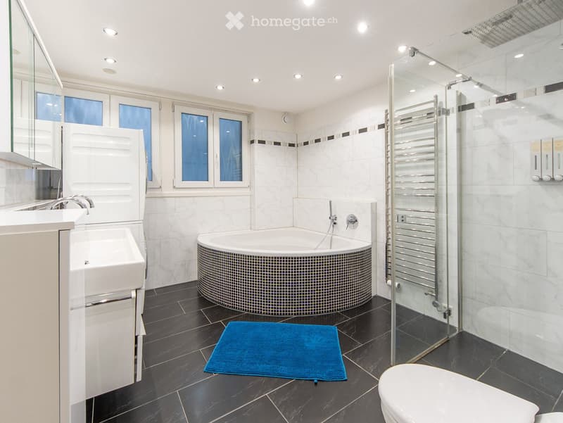 Modern bathroom with a round bathtub, white sink, toilet, large mirror, and a blue towel on the floor.