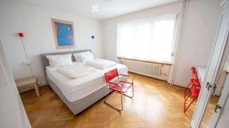 Bright bedroom with a double bed, two red chairs, a small table, and a picture frame on the wall. Natural light from a window illuminates the space.