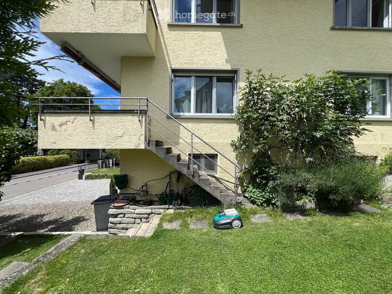 Front yard with a well-maintained lawn, a robotic lawnmower, a small staircase leading to a raised building entrance, and some greenery around the steps.