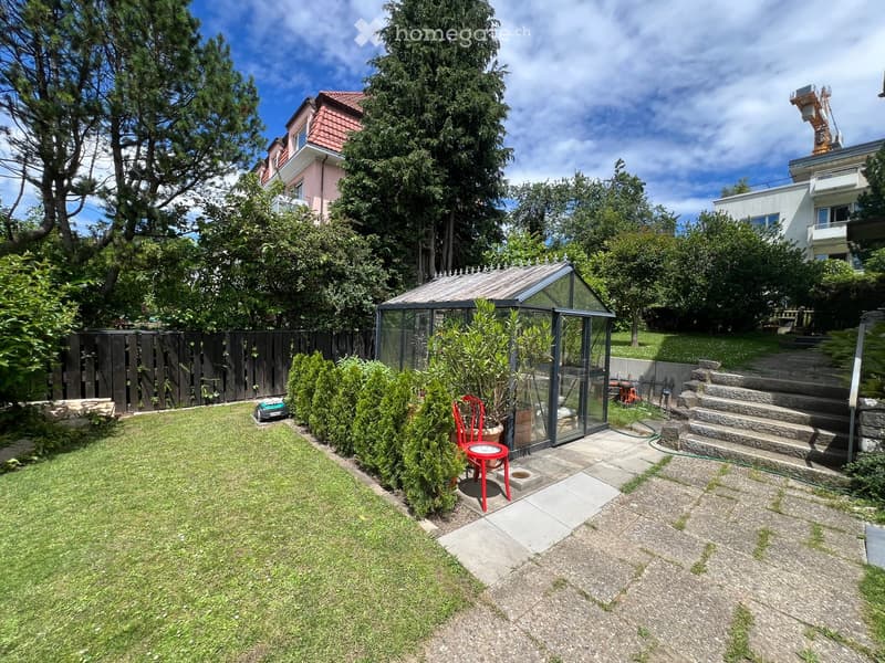 Well-maintained garden with a lawn, a small greenhouse, a red chair, and surrounding trees and shrubs.