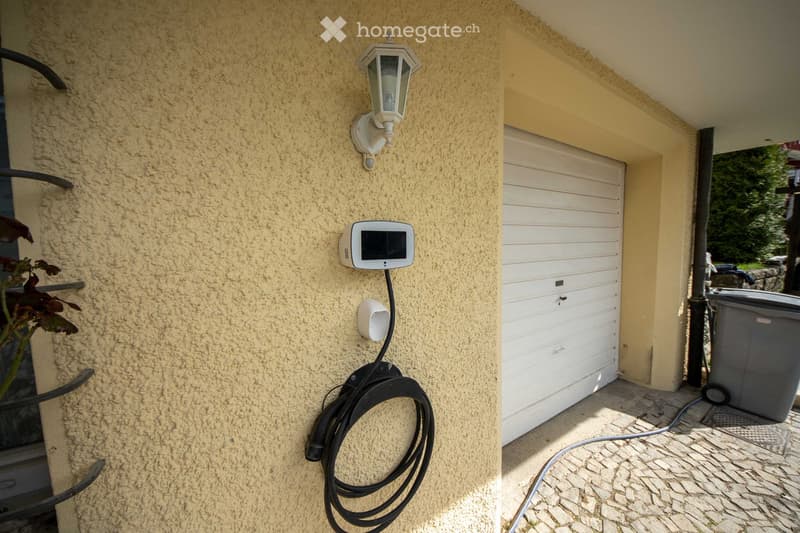 Exterior view of a house showing a textured beige wall with a mounted light fixture, a security camera, and a coiled black cable. A white garage door is visible to the right, with a paved stone pathway leading to it.