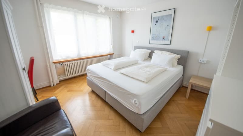 Cozy bedroom with a double bed, white bedding, a grey headboard, a black leather chair, a small wooden stool, a picture frame on the wall, and a floor lamp.
