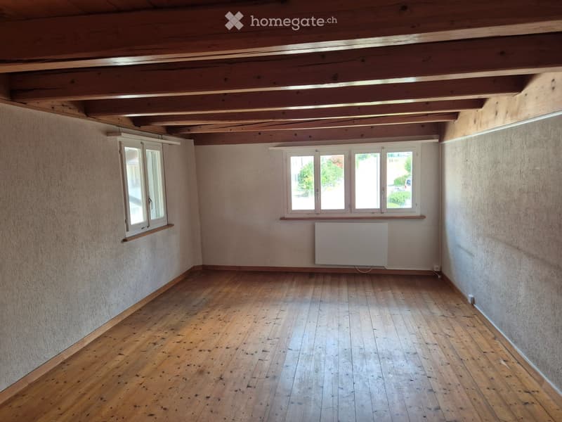 Empty room with wooden floor, exposed wooden ceiling beams, and two windows with white frames. A radiator is mounted on one wall.