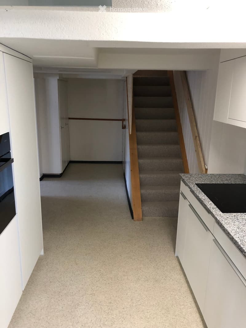 Empty hallway with carpeted floor, white walls, and a staircase leading upwards. Kitchen area visible on the right with white cabinets and a granite countertop.