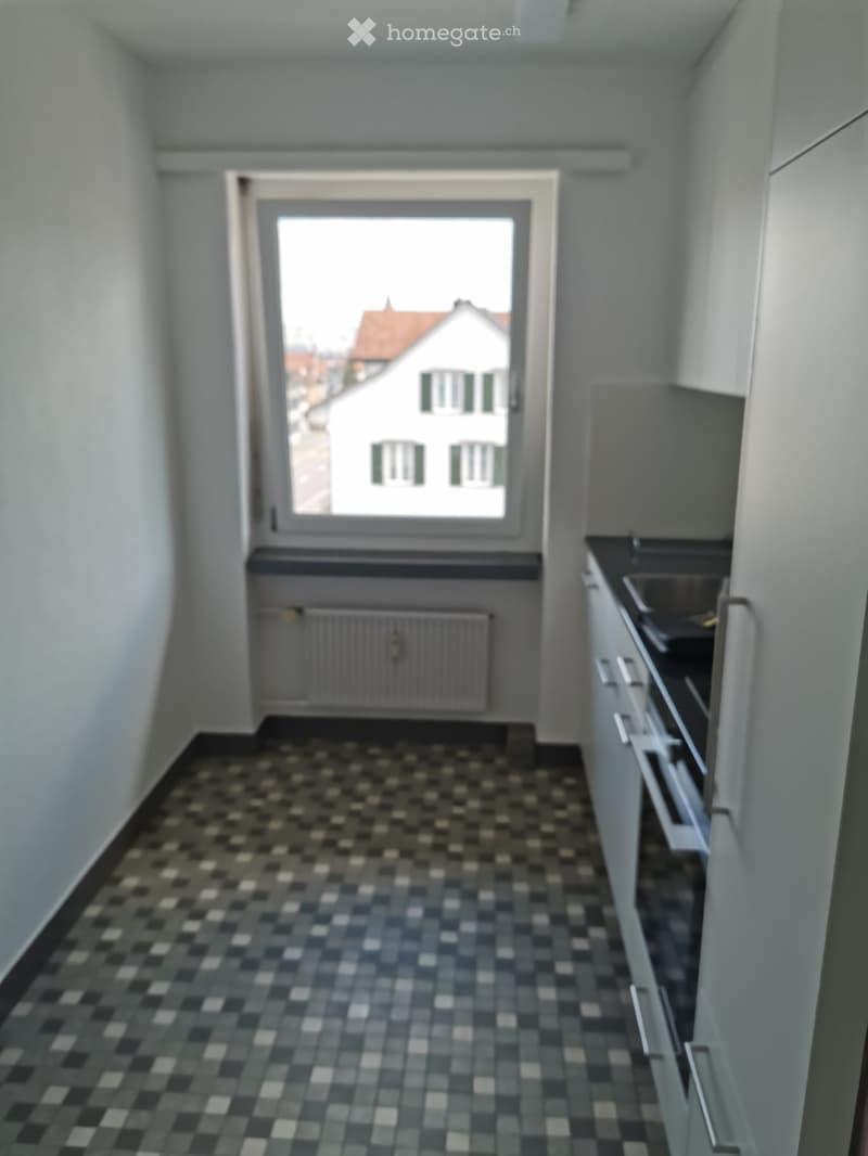 Empty kitchen, tiled floor, large window, modern appliances, radiator, open cabinet, sink, black countertop