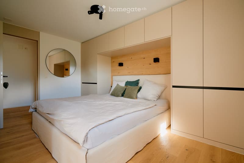 Modern bedroom with a neatly made bed, built-in wardrobe, and a round mirror on the wall.