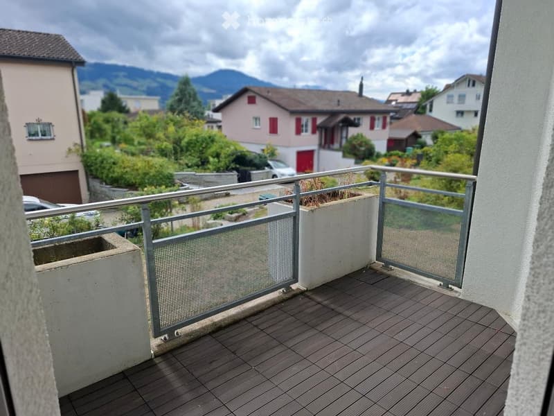 Balcony with wooden decking, metal railing, and potted plants, overlooking a residential area with houses and mountains.