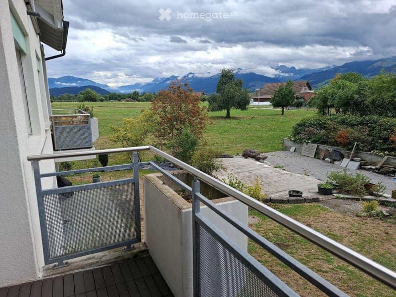 Balcony with metal railing overlooking a green field and distant mountains, with a small garden area below.
