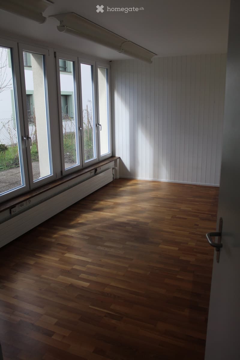 Empty room with wooden flooring, white paneled walls, large windows, and a radiator under the windows.