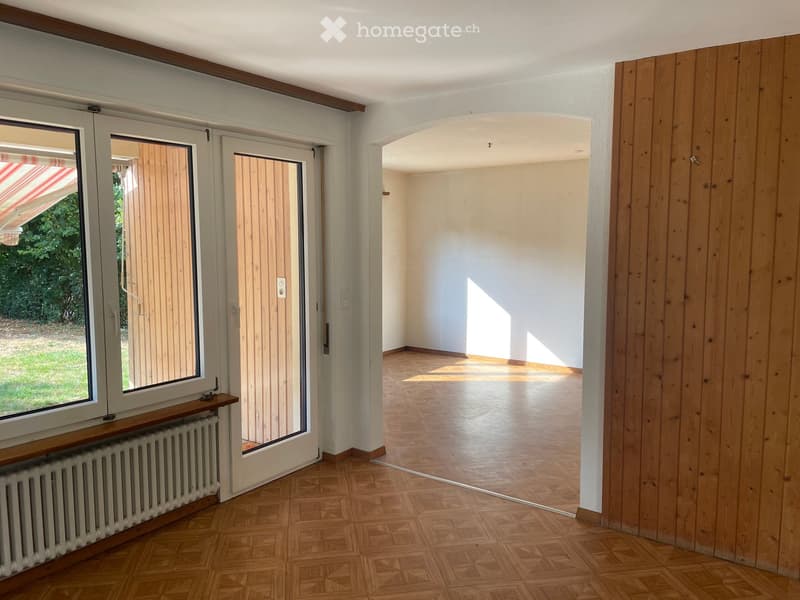 Empty room with wooden flooring, white walls, large window with blinds, and a radiator below the window. The room is connected to another empty space through an arched doorway.