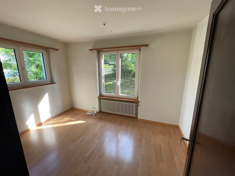 Empty room with wooden flooring, white walls, and large windows with wooden sills. A radiator is visible beneath one window.