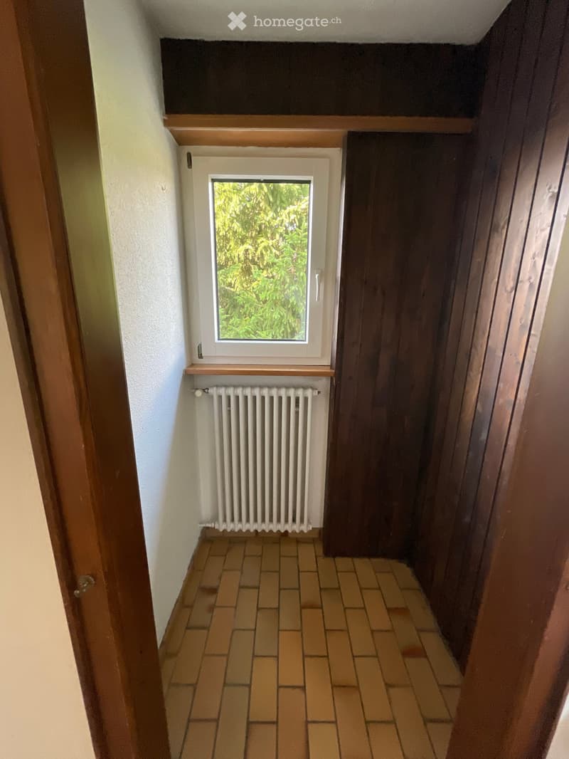 Empty hallway with tiled floor, white radiator, and a small window showing greenery outside.