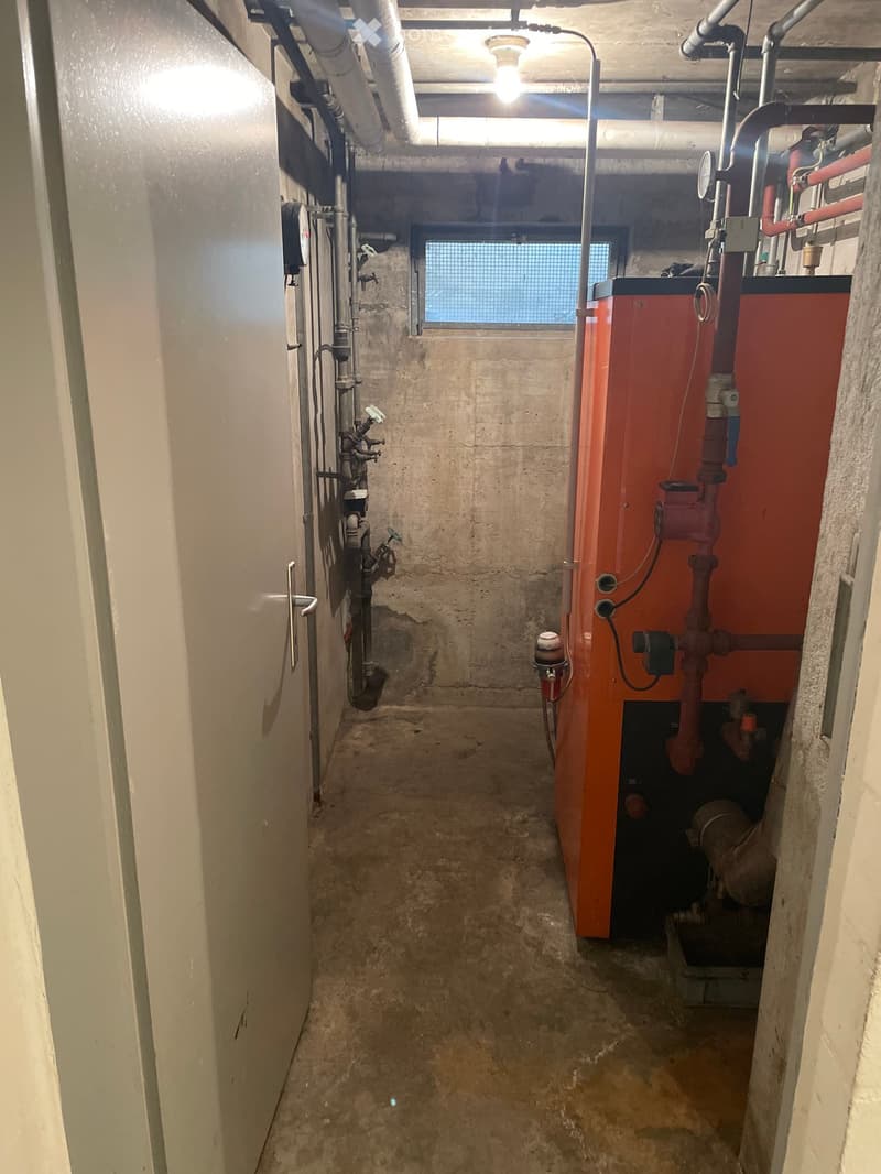 Utility corridor with exposed pipes, a red boiler unit, and a small window at the far end.