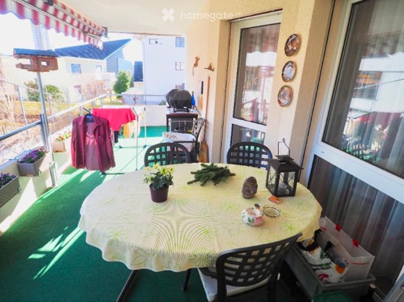 Balcony with green artificial turf, round table with floral tablecloth, chairs, decorative items, and a view of the neighborhood.