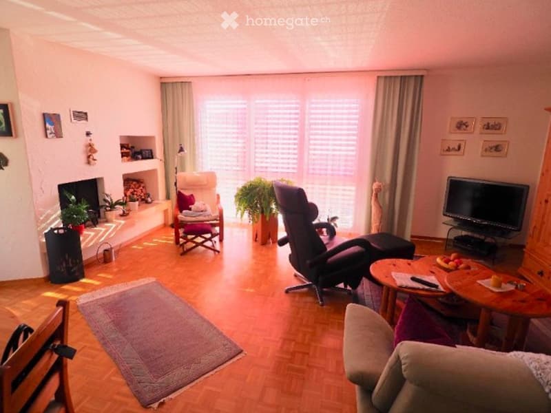 Bright living room with wooden flooring, fireplace, large windows with blinds, sofa, armchair, coffee table, TV, and decorative plants.