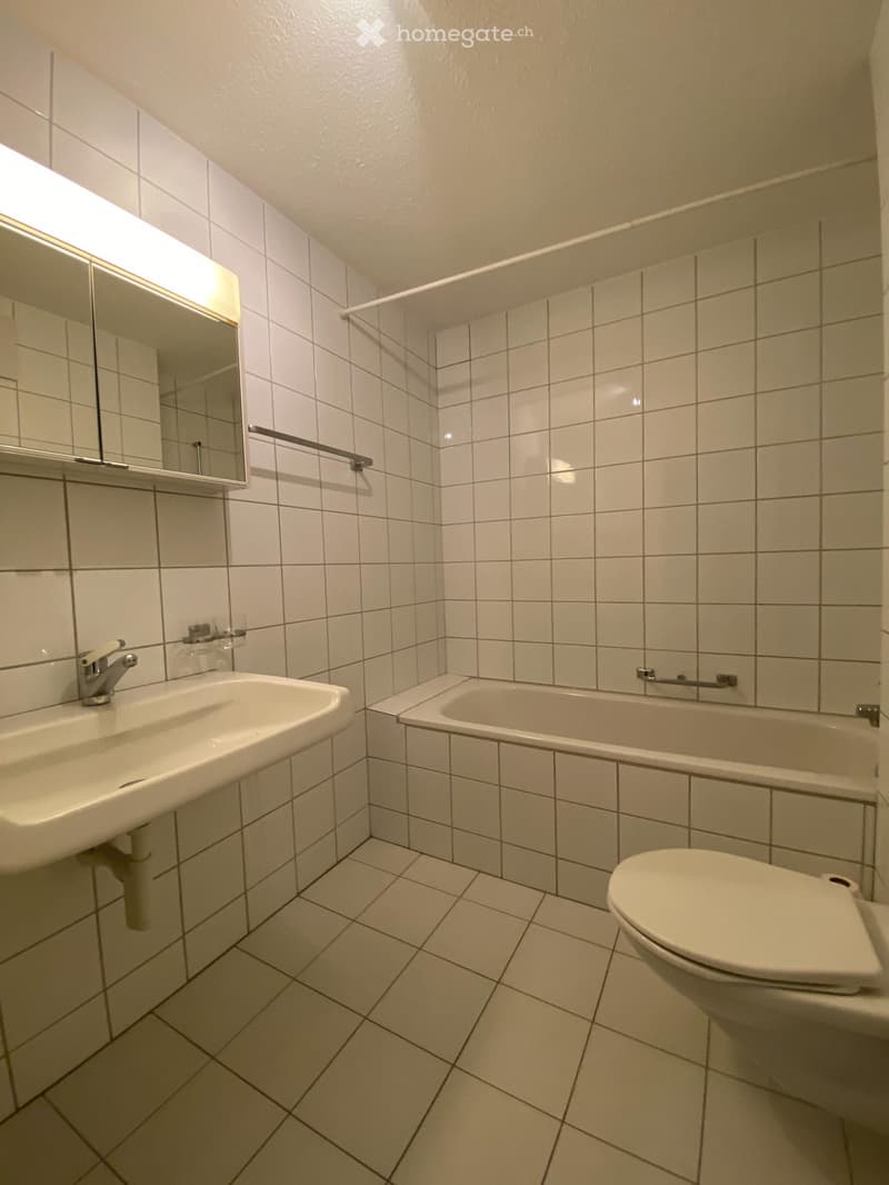 A small bathroom with white tiled walls and floor, featuring a sink with a faucet, a mirror above it, a bathtub with grab bars, and a toilet.