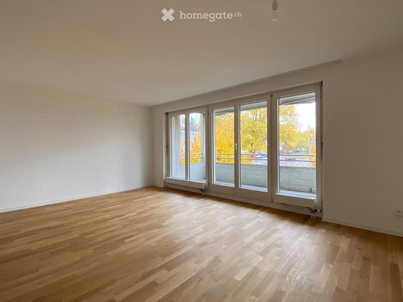 Empty room with wooden flooring and large windows, featuring white walls and radiators.
