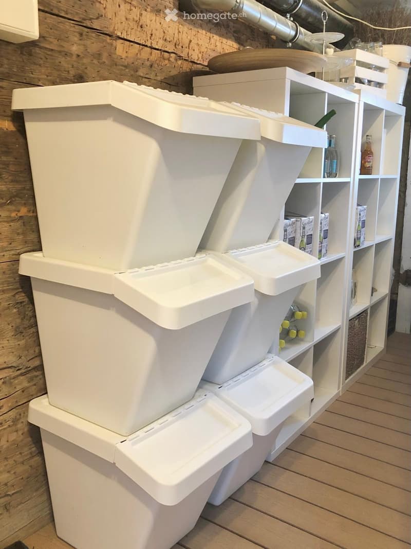 A room with white trash bins and shelves holding various items, including bottles and boxes, against a wooden wall.