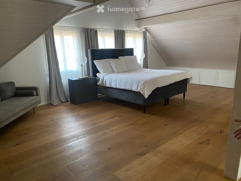 Cozy bedroom with wooden flooring, a large bed with white bedding, a nightstand with a lamp, and a grey armchair near the window.
