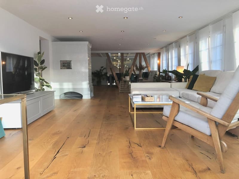 Modern living room with light wooden flooring, white walls, and large windows. Features a white sofa, glass coffee table, wooden armchair, TV stand, and decorative plants.