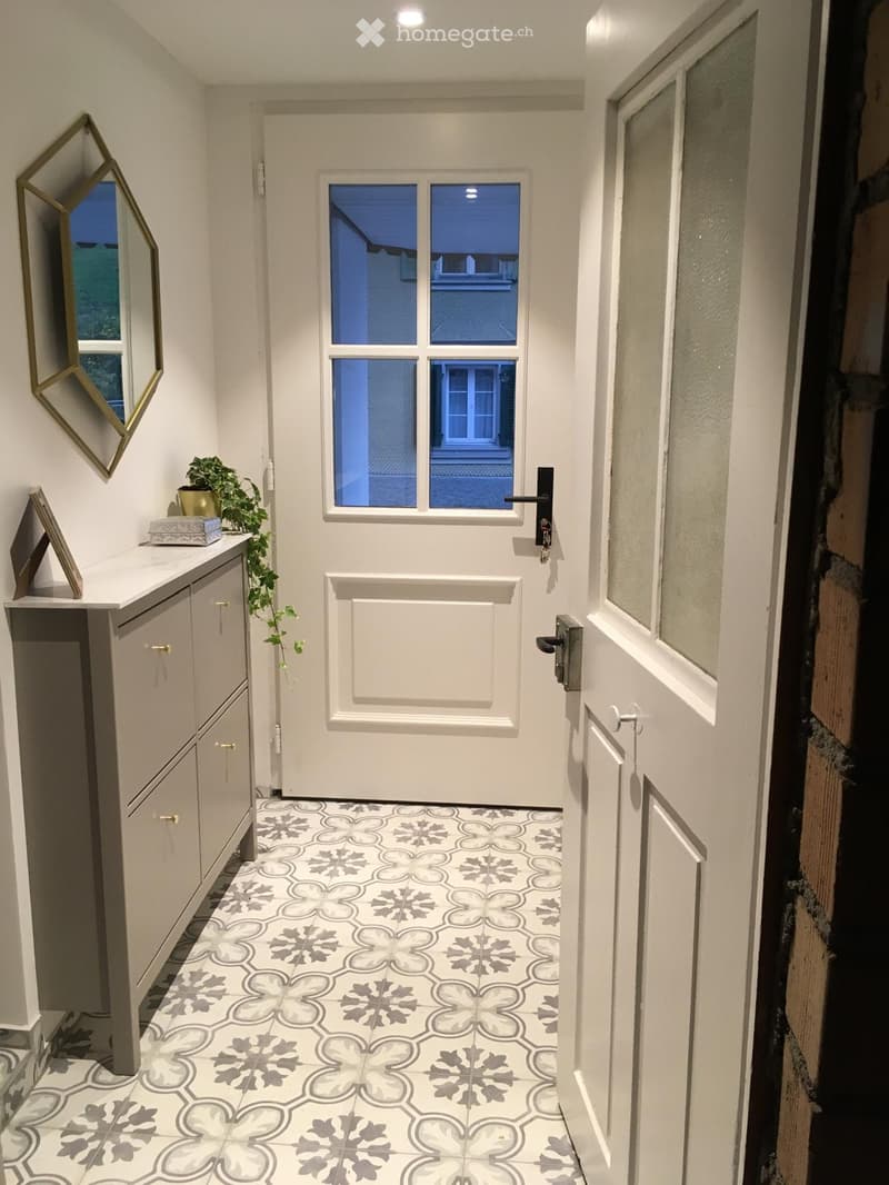 A narrow hallway with white walls, a decorative tiled floor, a small cabinet with a plant, and a mirror on the wall.