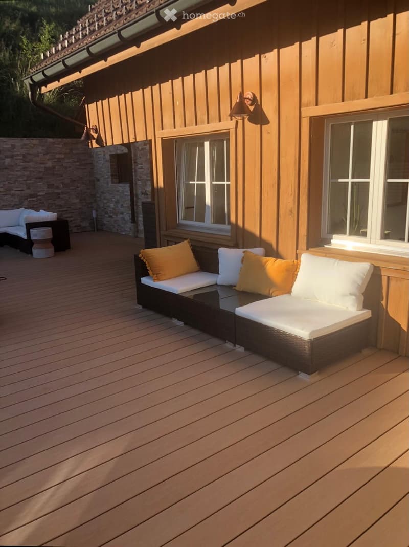 Wooden deck with a black sofa, white cushions, a small table, and a stool, adjacent to a stone wall and wooden house exterior.