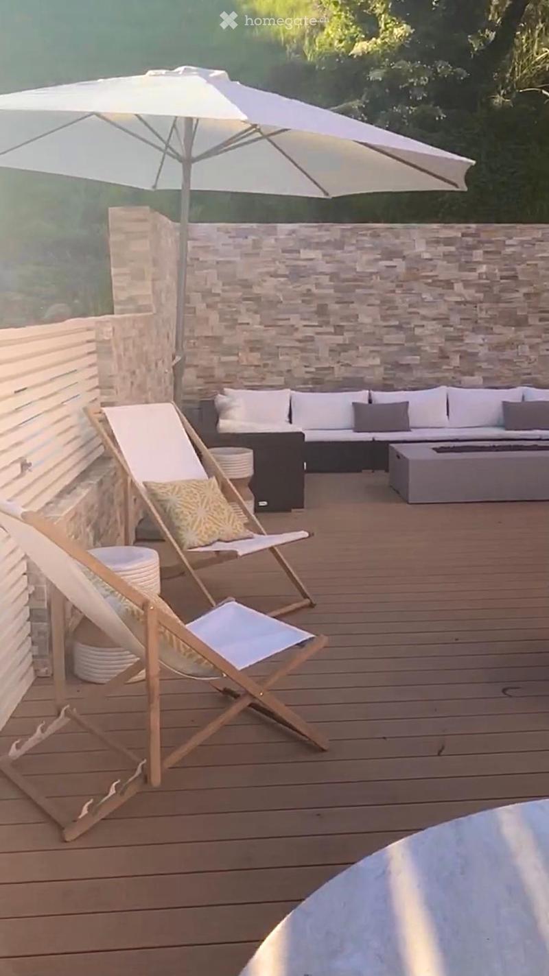 A modern outdoor terrace with wooden decking, a stone wall backdrop, a large umbrella, a sofa with cushions, a fireplace, and two wooden folding chairs.