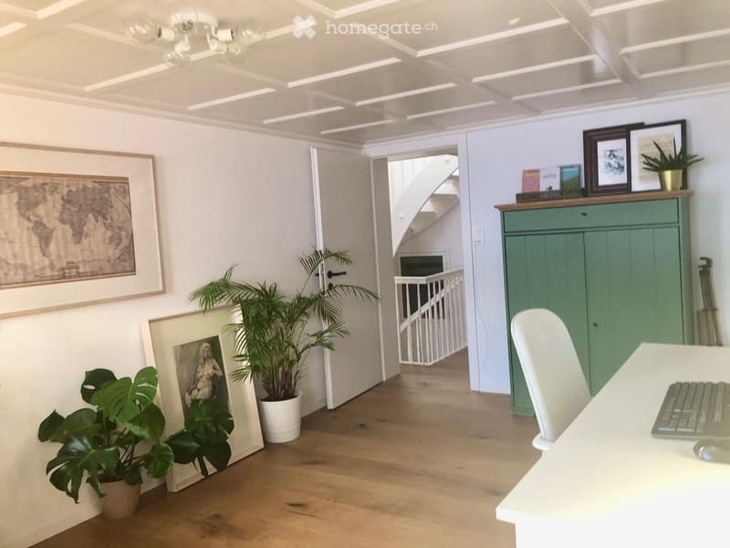 Bright, modern room with wooden flooring, white walls, and decorative elements including framed pictures and potted plants. A white chair and table with a laptop are positioned near a green cabinet, with a staircase leading upwards in the background.