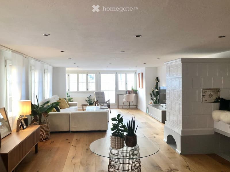 Bright living room with wooden flooring, large windows, a sofa, coffee table, armchair, table lamp, and a fireplace with a TV.