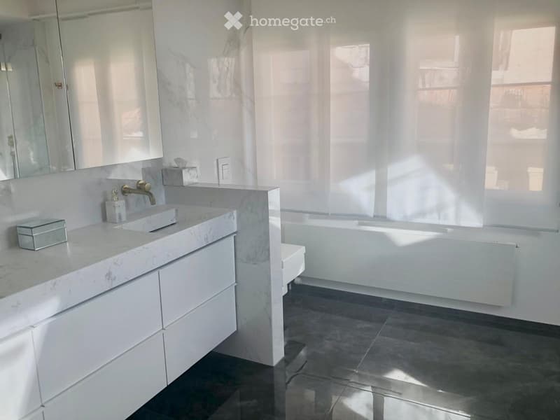Modern bathroom with a white marble sink, gold faucet, large mirror, white cabinets, bathtub, and toilet. Natural light streams in from a window with sheer curtains.