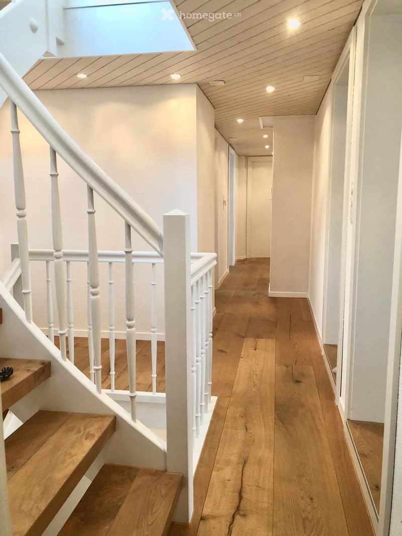 Spacious hallway with wooden flooring, white walls, and a staircase with white balustrade leading upwards. The ceiling has recessed lighting and a skylight.