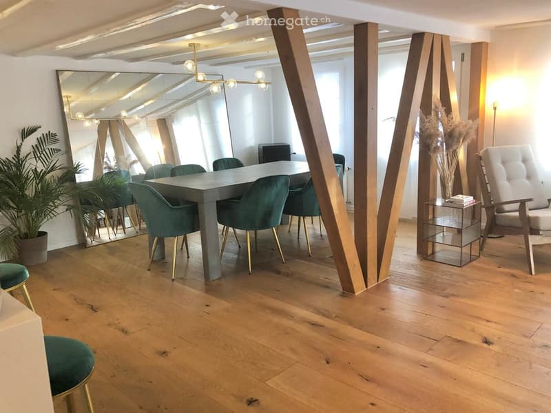 Modern, open-concept room with wooden flooring, a large grey table surrounded by green chairs, a white tufted chair, a side table with books, a large mirror, and decorative plants.