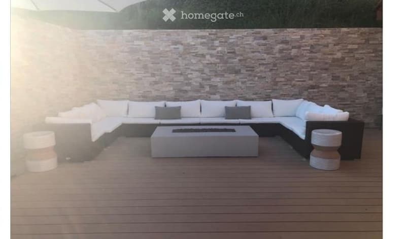 Modern outdoor seating area with a large sectional sofa, fireplace, and two white stools, set against a stone wall backdrop.