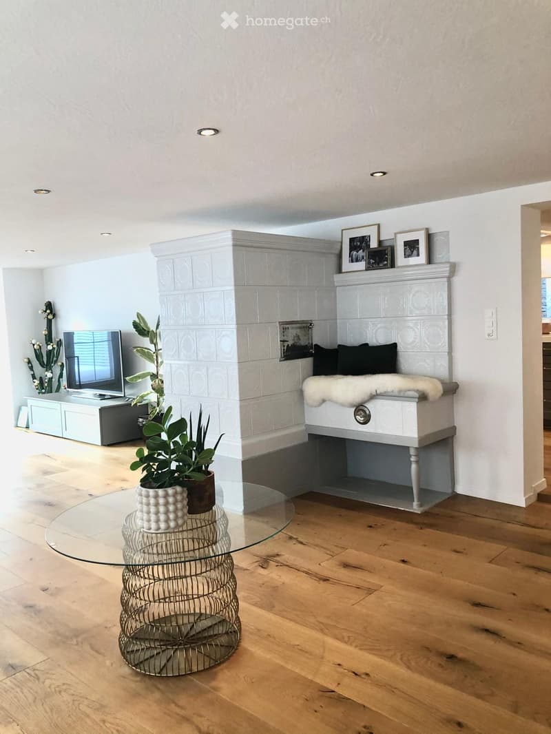 Modern living room with wooden flooring, a fireplace with a white fur throw, a glass coffee table, a television, and decorative plants.