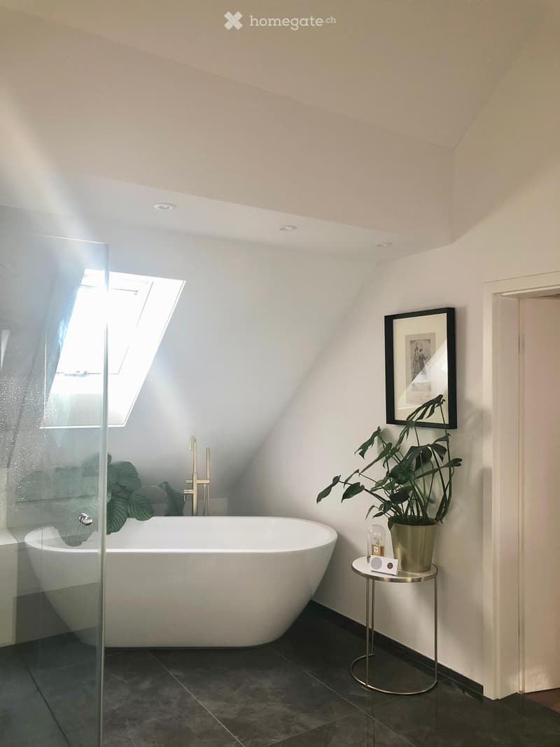 Modern bathroom with a freestanding bathtub, glass shower enclosure, sleek faucet, small round table with plant and speaker, and a framed picture on the wall.