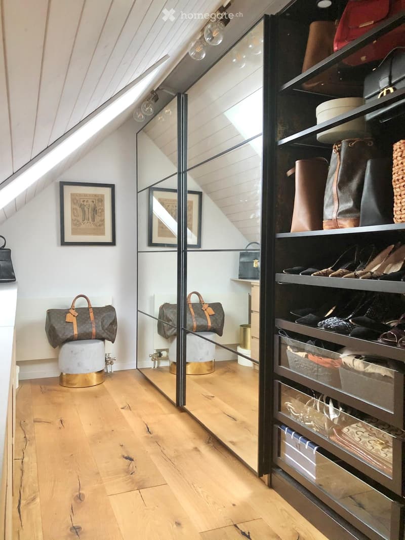 Attic room with wooden flooring, slanted ceiling, built-in shelving unit filled with bags and accessories, two framed pictures on the wall, and a small round stool with a handbag on top.