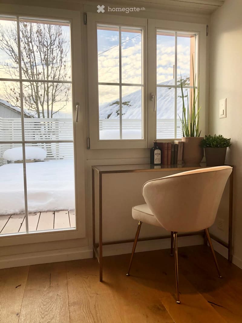 A modern, minimalist room with wooden flooring, large windows showing a snowy outdoor view, a sleek white chair with gold legs, a small table with books and a potted plant, and a bottle on the table.