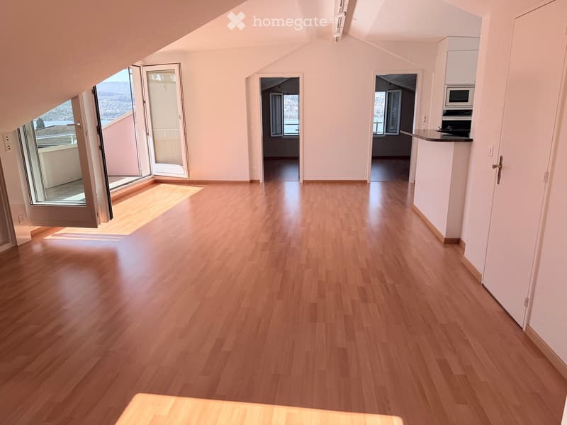 Bright, empty room with wooden flooring and white walls, featuring large windows and doors leading to a balcony with a scenic view.