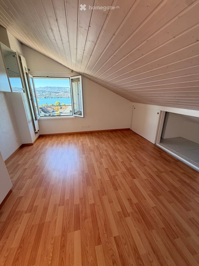 Empty attic room with wooden floors, slanted wooden ceiling, and large windows offering a scenic view of a lake and mountains.