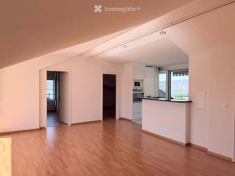 Bright, open-plan living and dining area with wooden flooring, white walls, and modern kitchen appliances including a refrigerator, microwave, and oven. A large window offers a scenic view.