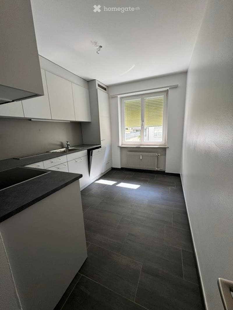 Empty kitchen with white cabinets, dark tiled floor, and a window with blinds. No furniture or appliances are present.
