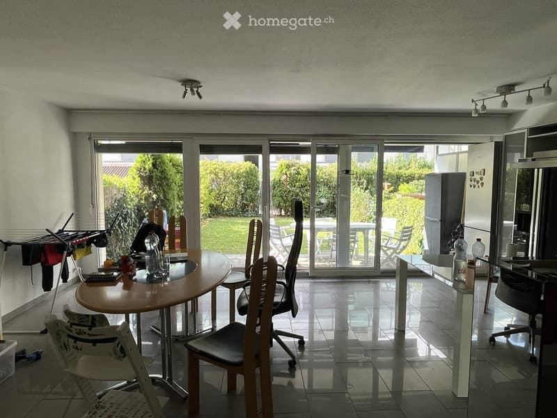 Dining area with a round wooden table, several chairs, and a view of a garden through large sliding glass doors. There is also a refrigerator and a mirror visible.