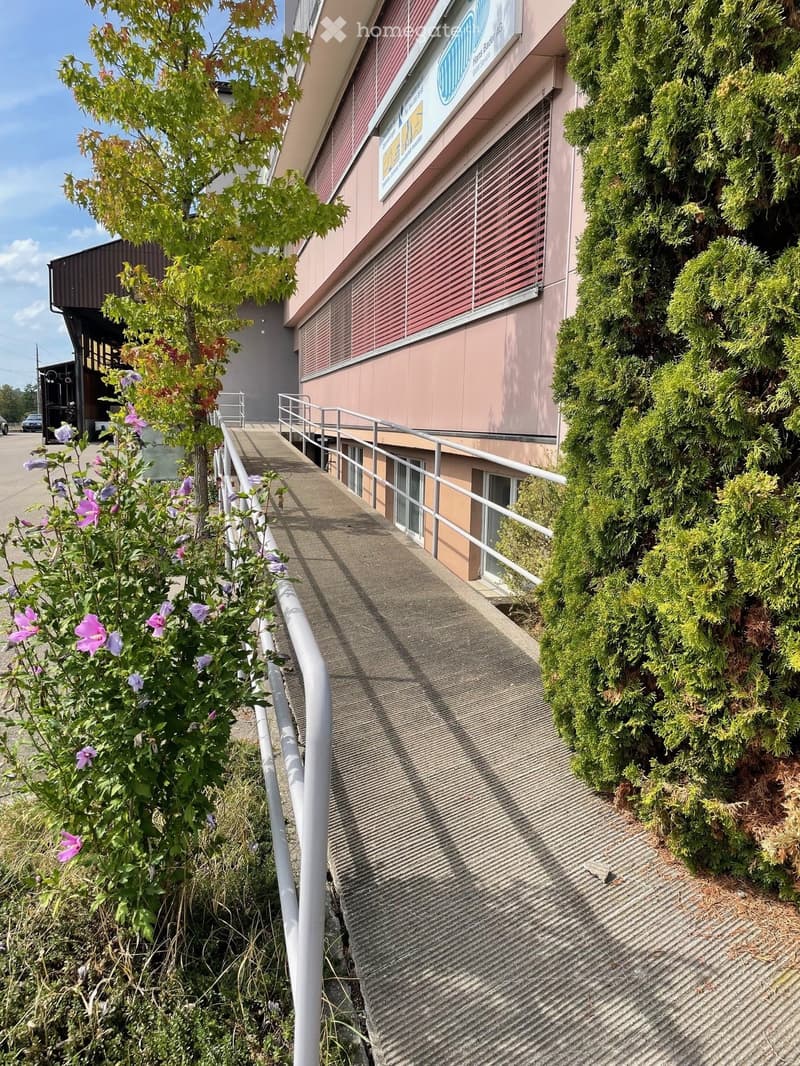 Exterior view of a two-story building with a ramp leading to the entrance, surrounded by greenery and flowers.