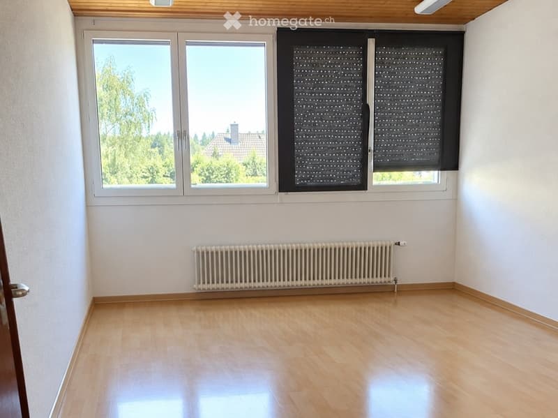Empty room with wooden flooring, white walls, large windows with blinds, and a radiator.
