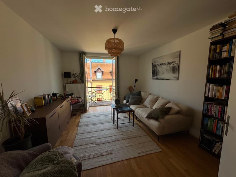 Modern living room with a white sofa, coffee table, bookshelf, and a balcony view. Features include wooden flooring, a woven pendant light, and a cozy seating area.