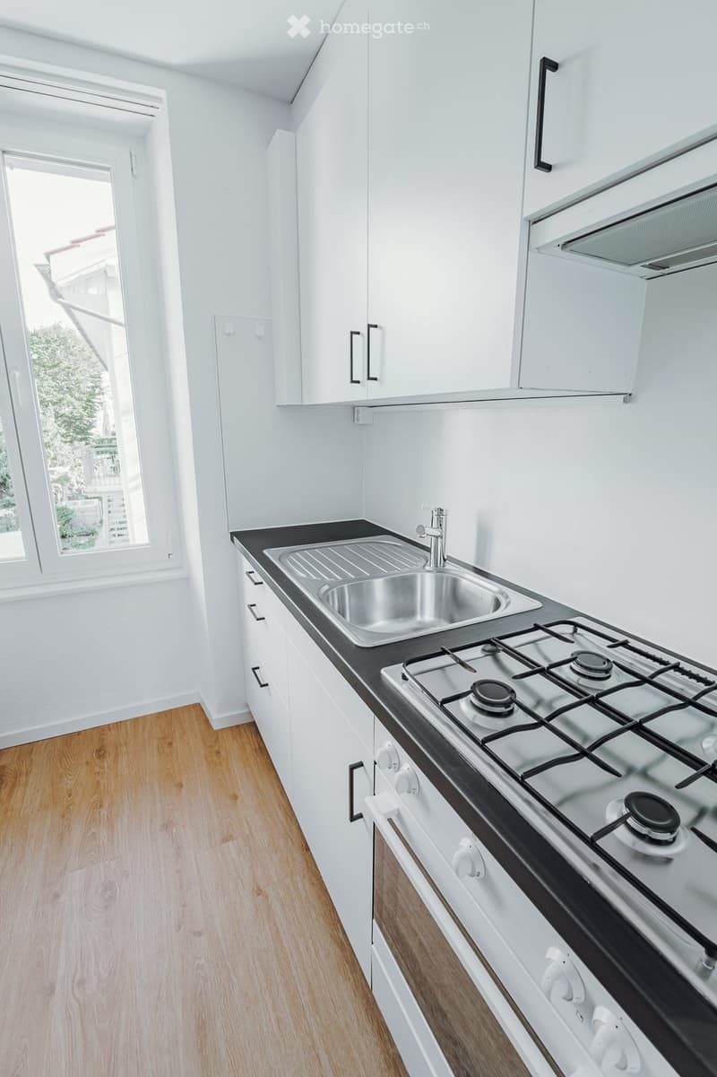 Modern kitchen with white cabinets, a stainless steel sink, and a gas stove. The floor is wooden, and there is a window providing natural light.