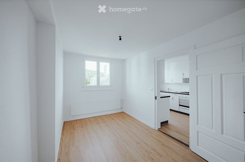 Empty room with white walls, wooden flooring, and a window. Adjacent kitchen area with white cabinets and appliances is visible through an open doorway.
