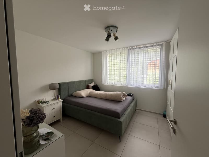 A modern bedroom with a grey upholstered bed, nightstand, lamp, and decorative vase on a small table. The room features tiled flooring and large windows with blinds.