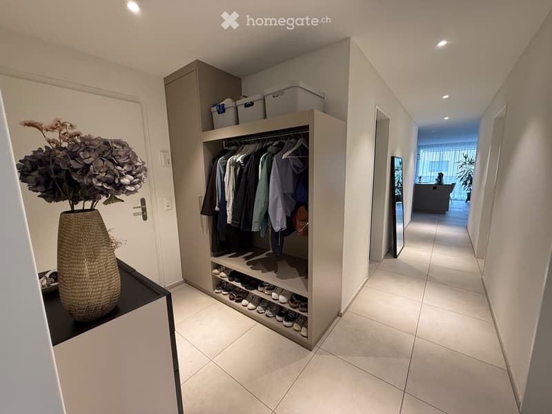 Modern hallway with tiled flooring, built-in closet with clothing and shoes, vase with flowers on a small table, and a view into an adjacent room with a kitchen area.